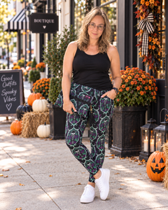 A woman stands on a storefront sidewalk wearing patterned pants with a black tank top and white sneakers. The pants have a dark base with a repeating green and purple witch hat motif.