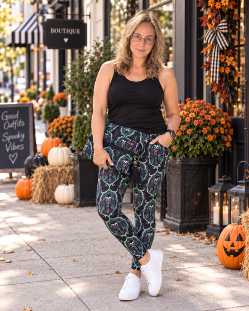 A woman stands on a storefront sidewalk wearing patterned pants with a black tank top and white sneakers. The pants have a dark base with a repeating green and purple witch hat motif.
