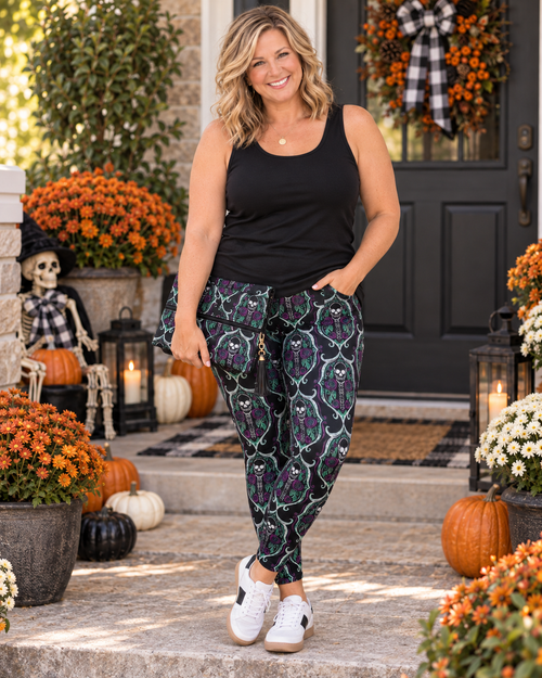 A woman stands on a front porch decorated for Halloween, wearing a black tank top and patterned jogger pants with white sneakers. The pants feature a dark green trellis pattern with purple and orange floral motifs.