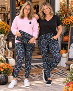 Two women modeling Halloween-themed leggings with a purple and black spiderweb and ghost pattern, standing in front of a decorated storefront.