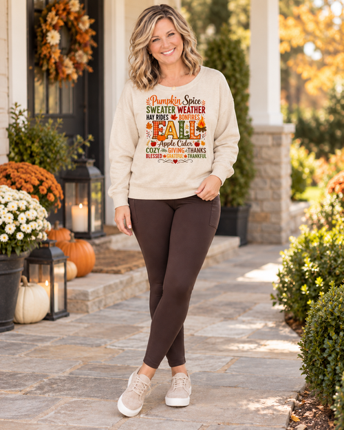 A woman stands on a stone walkway in front of a decorated autumn porch, wearing a cream sweatshirt with a colorful 'Pumpkin Spice' fall graphic and dark brown leggings.