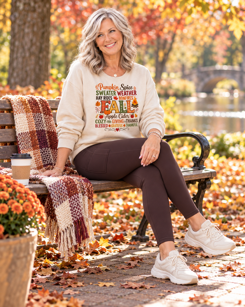 A woman sits on a wooden park bench surrounded by autumn leaves, wearing a cream-colored sweatshirt with a colorful fall-themed graphic that reads 'Pumpkin Spice SWEATER WEATHER MY FEELINGS FALL Apple Cider COFFEE CHIPS BLESSED BEYOND SUMMER.'.