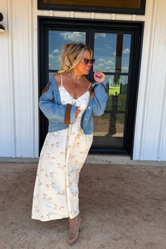 A woman stands outdoors in front of a white building with a black-framed window, wearing a long cream maxi dress with a small floral print, a light blue denim jacket with brown shoulder patches, and brown ankle boots.