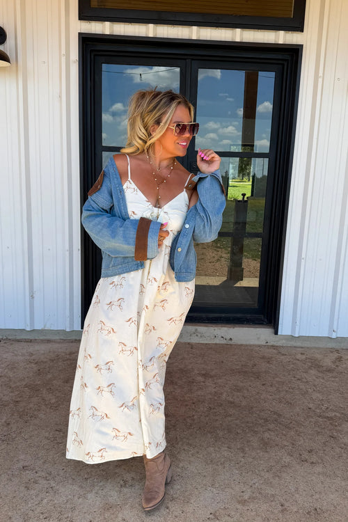 A woman stands outdoors in front of a white building with a black-framed window, wearing a long cream maxi dress with a small floral print, a light blue denim jacket with brown shoulder patches, and brown ankle boots.