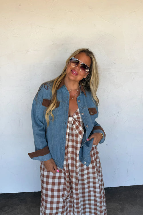 A woman posing in a light blue denim jacket with brown leather-like patch pockets over a brown and white gingham dress, wearing sunglasses against a white stucco wall.