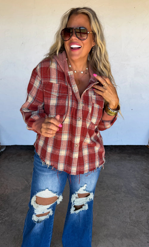 A woman wearing a red and beige plaid hooded button-up shirt, blue ripped jeans, and sunglasses, posing in front of a plain light wall.