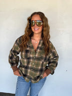 A woman with long wavy brown hair wearing a green and brown plaid button-down shirt, blue jeans, and reflective aviator sunglasses, standing against a light-colored stucco wall.