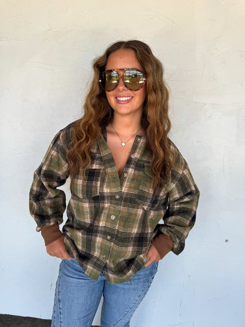 A woman with long wavy brown hair wearing a green and brown plaid button-down shirt, blue jeans, and reflective aviator sunglasses, standing against a light-colored stucco wall.