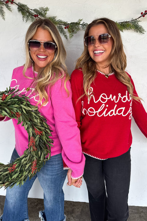 Two women wearing festive sweaters with holiday-themed text, standing against a white wall with greenery.