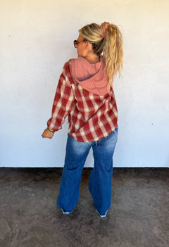 A woman stands with her back to the camera wearing a red and white plaid hooded flannel shirt over a dusty pink hoodie, blue flare jeans, and sunglasses, set against a plain white wall and concrete floor.