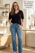Woman wearing a black shirt and blue jeans standing in a bright room with wooden furniture and plants.