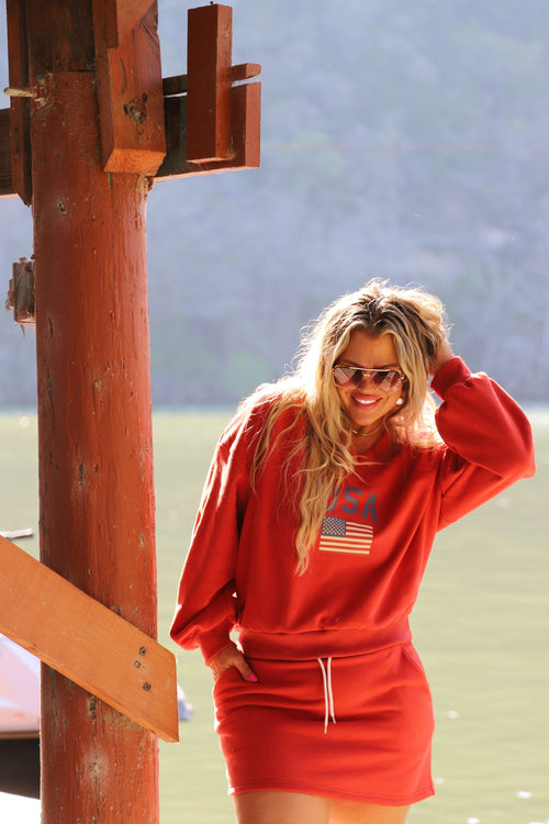 Model wearing an oversized red V-neck pullover with a "USA" flag graphic on the chest, standing on a sunny dock.