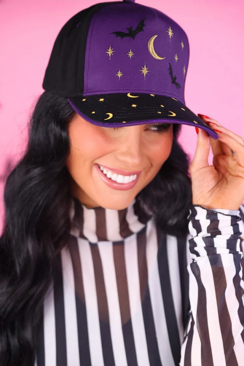 A woman wearing a purple and black baseball cap with embroidered bats, crescent moon, and stars, posed against a solid pink background.