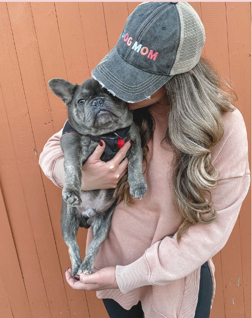 A woman wearing a gray 'DOG MOM' trucker hat and a dusty pink sweater holds a gray French Bulldog against a brown wooden fence background.