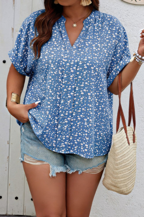Woman wearing a blue floral blouse and denim shorts, holding a straw bag against a white wall.