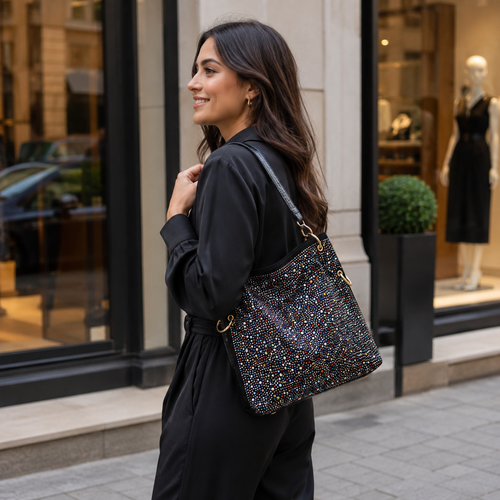 A woman in a black outfit carries a large, dark rhinestone covered shoulder bag with gold hardware and a black strap, walking on a city street in front of a storefront.