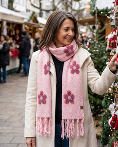 A woman at an outdoor Christmas market wearing a light pink knit scarf with darker pink flower motifs and fringed ends, paired with a cream coat.