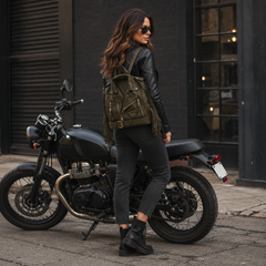 A woman wearing a black leather jacket, black jeans, and black boots stands next to a black motorcycle, wearing an olive green fringed backpack. She is photographed in an urban street setting with a dark brick building in the background.