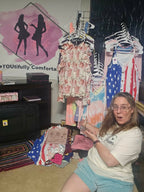 Enthusiastic boutique owner pointing to racks of colorful floral tops, patriotic tanks, and tie-dye dresses for a clearance event.