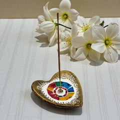 Heart-shaped incense holder with colorful design on a white surface with white flowers in the background.