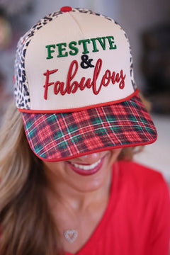 A woman wearing a festive trucker hat with a cream front panel, leopard print side panels, a red plaid visor, and embroidered text reading 'FESTIVE & Fabulous'.