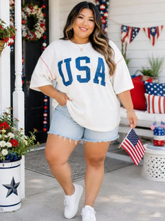 A woman stands on a decorated front porch wearing an oversized white USA graphic T-shirt with blue lettering, light denim shorts, and white sneakers, while holding a small American flag.
