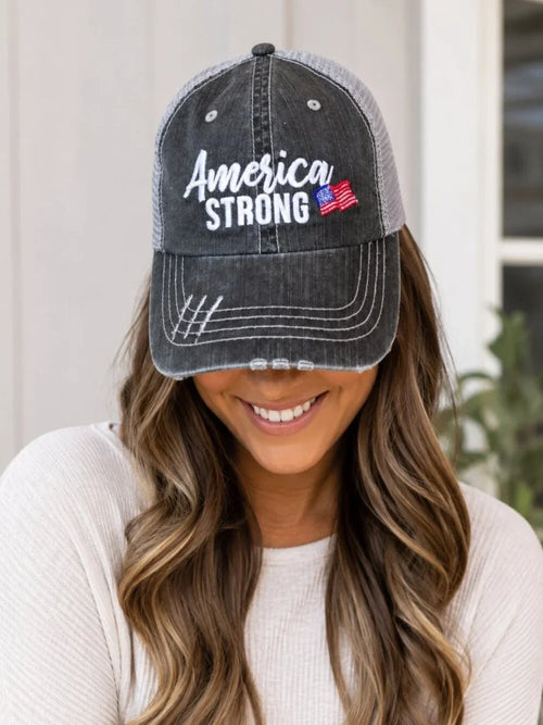 A woman wearing a dark gray washed trucker hat with 'America Strong' embroidered on the front, featuring a distressed brim and mesh back.