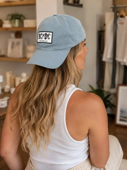 A woman with long blonde hair is shown from behind wearing a light blue baseball cap with a black and white graphic patch that reads ACDC. She is wearing a white ribbed tank top in what appears to be a boutique setting.
