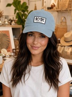 A woman wearing a light blue AC/DC baseball cap with a black-and-white embroidered logo patch on the front, photographed in a warm, styled indoor boutique setting.