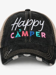 A distressed black trucker cap with 'Happy CAMPER' embroidered on the front. The front panel features a washed black denim-like texture with white, pink, blue, and green embroidery, while the mesh back is a natural tan/beige color.
