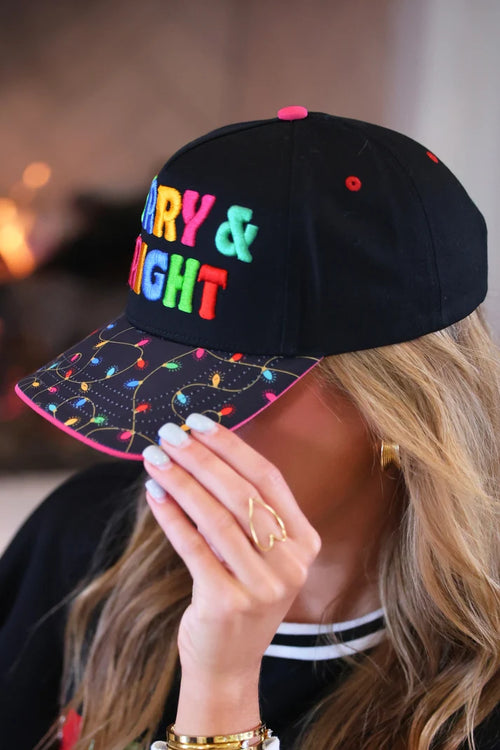 A woman wearing a black baseball cap with colorful embroidered text reading 'MERRY & BRIGHT' on the front and multicolored Christmas string lights printed along the brim. The photo is cropped closely on the model's face, hand, and hair, with a softly blurred indoor background.
