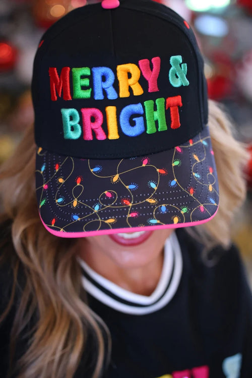 A person wearing a black baseball cap with colorful embroidered 'MERRY & BRIGHT' text and a brim printed with multicolored Christmas string lights, photographed in a festive indoor setting.