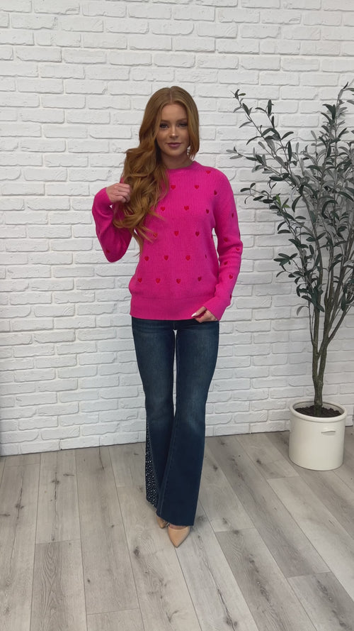 Model wearing the Eat Your Heart Out Pink Heart Sweater with jeans, showcasing embroidered heart details.