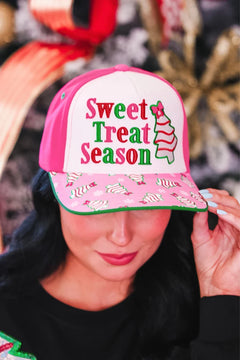 A woman wearing a pink and white trucker-style baseball cap with 'Sweet Treat Season' embroidered on the front and a pink brim covered in small candy cane prints.