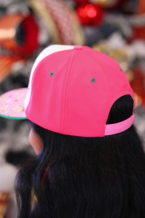 A close-up back view of a person wearing a bright pink and white baseball cap with a pink visor and green embroidered eyelets, shown against a blurred festive background with red ornaments.