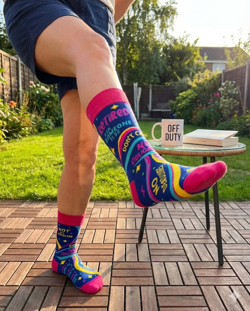 A person kicking their leg up outdoors wearing vibrant blue and pink novelty crew socks that read "Retired Ask Someone Else" and "No Deadlines".